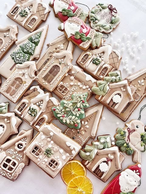 Decorated gingerbread houses and Christmas cookies with green frosting, ribbons, and dried orange slices arranged on white surface